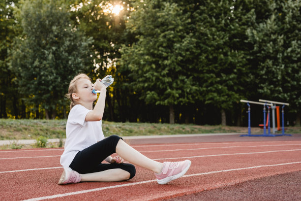 小女孩运动后坐在跑道上喝水的照片。儿童健康积极的生活方式理念。图片下载