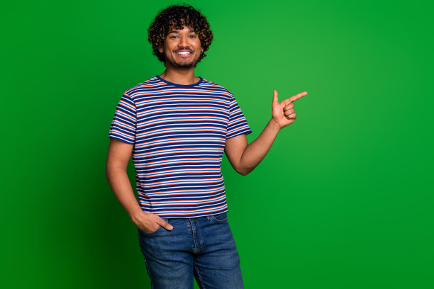 照片肖像年轻的印度学生有趣的家伙波浪的头发，穿条纹t恤食指他的宣传孤立的绿色背景图片下载