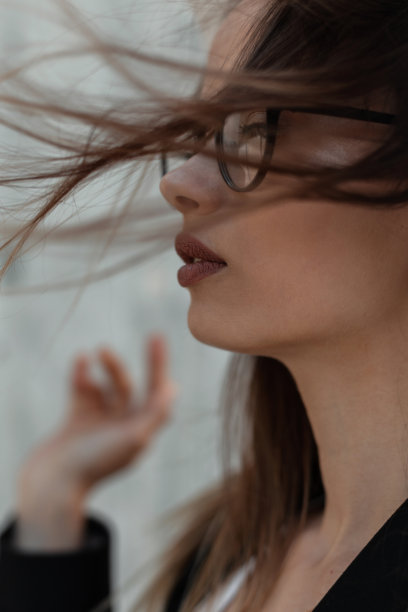 特写照片女性美丽的脸，漂亮的嘴唇在时尚的眼镜，时尚的黑色衣服在灰色的背景，在刮风的日子户外。图片下载
