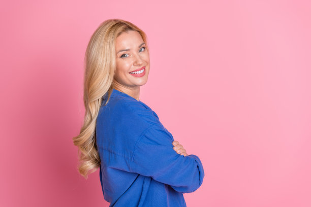 侧面侧面照片满意的女人波浪头发穿着蓝色衬衫抱臂交叉牙齿微笑孤立的粉红色背景图片下载