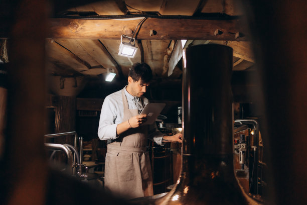 啤酒质量控制。戴着围裙和防护面具的男子拿着数码平板电脑看着啤酒厂的啤酒杯，自由的空间图片下载