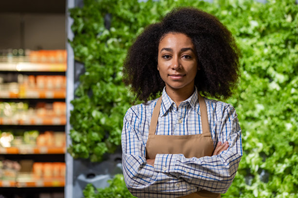 一位自信的女超市工作人员双臂交叉站在农产品展示区的生菜前。她面带微笑，穿着制服显得专业而自信图片下载