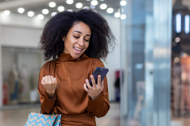 微笑的年轻非洲裔美国妇女站在商场里，拿着纸袋，用着手机，开心地看着屏幕，用手做出胜利的手势图片下载