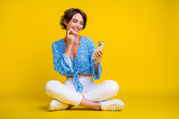 全尺寸照片，聪明可爱的女人穿着蓝色上衣，坐在看智能手机，手指放在嘴唇上，孤立在黄色背景上图片下载