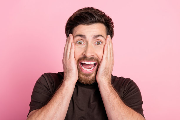 照片上的男人惊讶得说不出话来，深色头发的胡茬，穿着棕色t恤，双手放在脸颊上，孤立在粉红色的背景上图片下载