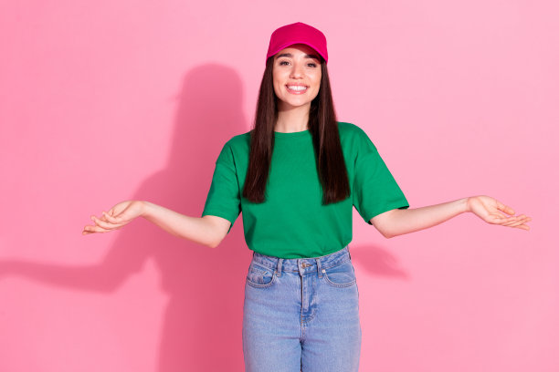 照片中迷人积极的女人穿着绿色t恤，粉红色的帽子，对比手臂空白的空间，孤立的粉红色背景图片下载
