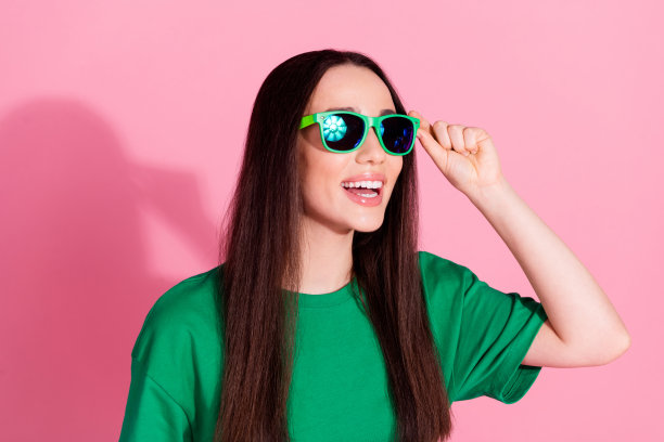 照片中迷人积极的女人穿着绿色t恤，墨镜，显示出两个v字的空白空间孤立的粉红色背景图片下载