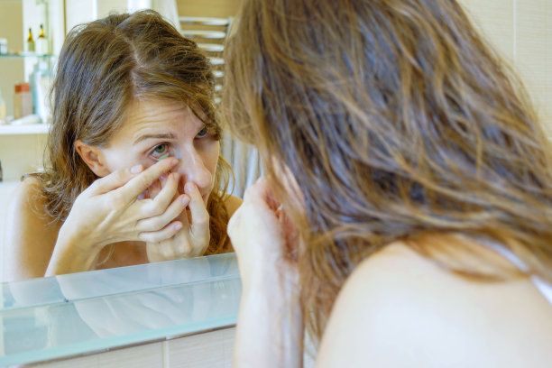 一名年轻女子站在浴室的镜子前，将一副绿色隐形眼镜插入眼睛——这是一张特写照片。图片下载