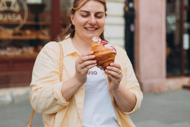 30多岁的漂亮超重黑发女孩在街上摆姿势，她手里拿着大羊角面包。不健康的肥胖食品，高热量的零食，边走边吃，外卖。图片下载