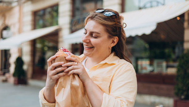 30多岁的漂亮超重黑发女孩在街上摆姿势，她手里拿着大羊角面包。不健康的肥胖食品，高热量的零食，边走边吃，外卖。图片下载