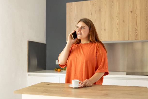 在慵懒的早晨，在舒适的厨房里，悠闲随意的年轻美丽的快乐微笑的女人喝着咖啡。30多岁的女人在家里打电话。高质量照片图片下载