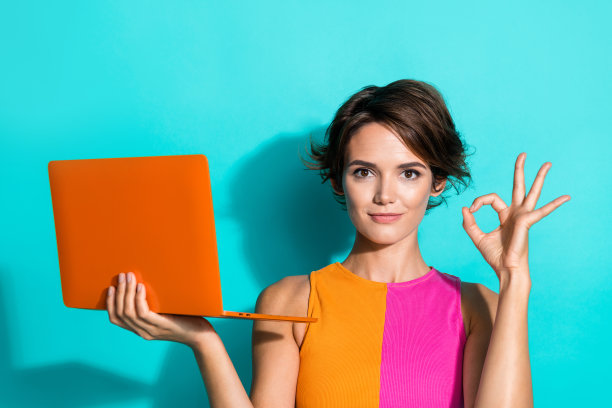 照片中，一个冷静自信的女人穿着粉色的橙色上衣，展示着ok的标志，发短信，现代小工具孤立的蓝绿色背景图片下载