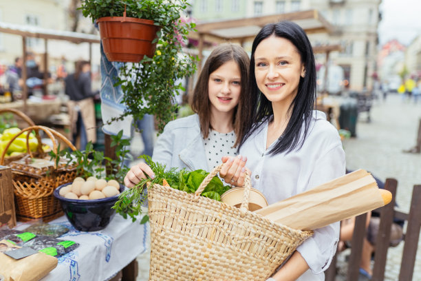 在一个阳光明媚的早晨，两姐妹在农贸市场的有机蔬菜摊位上买菜，年轻女子提着一个篮子。销售，购物，生态健康食品和人的理念。图片下载