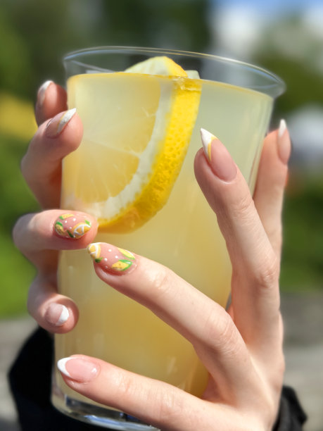 特写图片中无法辨认的女性双手握着浑浊的、老式的、装在塑料杯里的柠檬水饮料，展示了柠檬水果和柑橘片的指甲油美甲设计，阳光明媚的户外环境背景，重点放在前景上图片下载