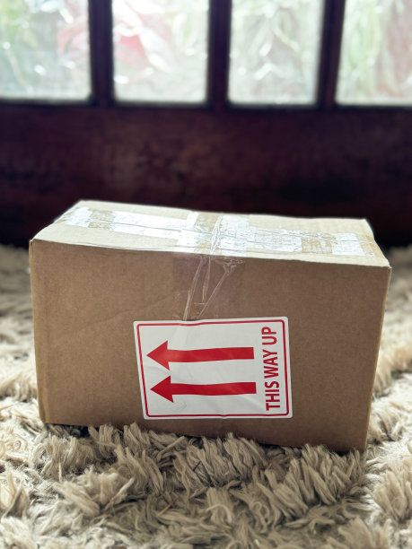 特写图片:快递包裹贴在前门垫子上的方向错误，带有红色方向箭头指示标签的“This way Up”纸板箱，带有玻璃板背景的木制前门，蓬松的门垫，聚焦于前景，图片下载
