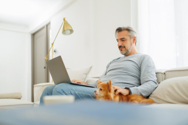 在舒适的客厅里，一个面带微笑的中年男人一边摆弄着笔记本电脑，一边抚摸着沙发上的小吉娃娃狗，表现出专注而轻松的心情。图片下载