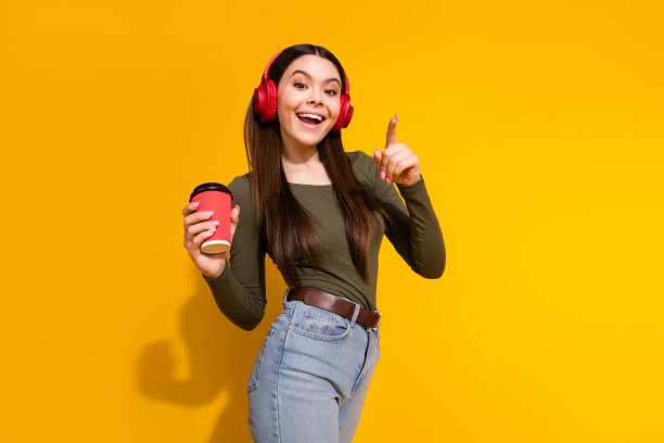 漂亮少女的照片肖像，耳机，咖啡饮料，手指，穿着时髦的卡其布服装，孤立在黄色背景上图片下载