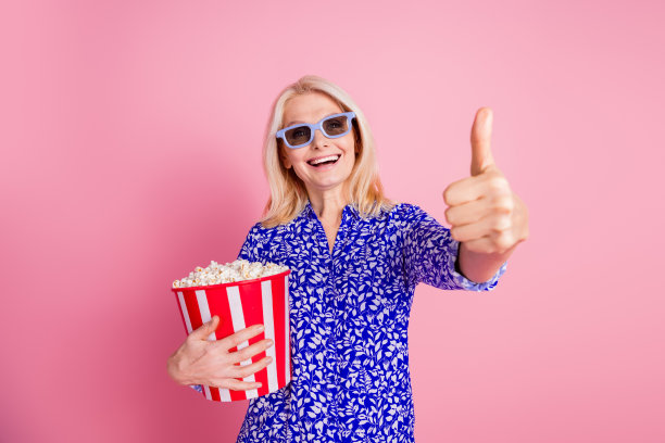 照片漂亮的老太太3d眼镜电影院爆米花拇指向上穿蓝色衬衫孤立在粉红色的背景图片下载