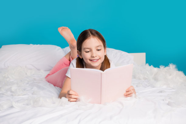 漂亮可爱的女孩躺在舒适的床上阅读孤立的蓝色背景的照片图片下载