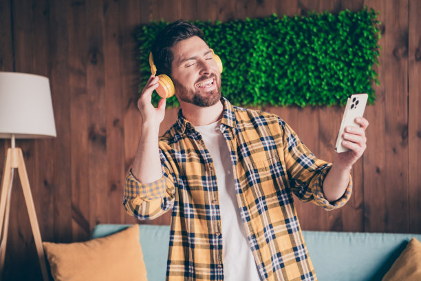 照片开朗积极的家伙穿着格子衬衫，耳机听音乐，现代设备在室内的房子公寓房间图片下载