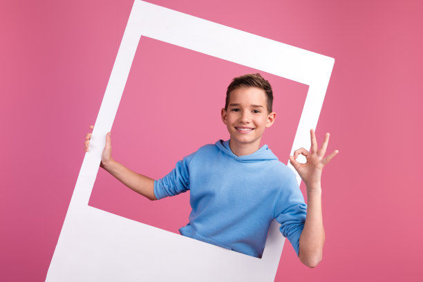 可爱的十几岁男孩的照片肖像拿着即时相框ok批准穿着时尚的蓝色衣服孤立在粉红色的背景图片下载