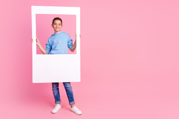 可爱的十几岁男孩的全身照片拿着即时相框，穿着时髦的蓝色衣服，孤立在粉红色的背景上图片下载