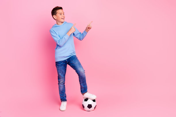 全身照片可爱的十几岁的男孩踢足球点看空穿着时尚的蓝色衣服孤立在粉红色的背景图片下载