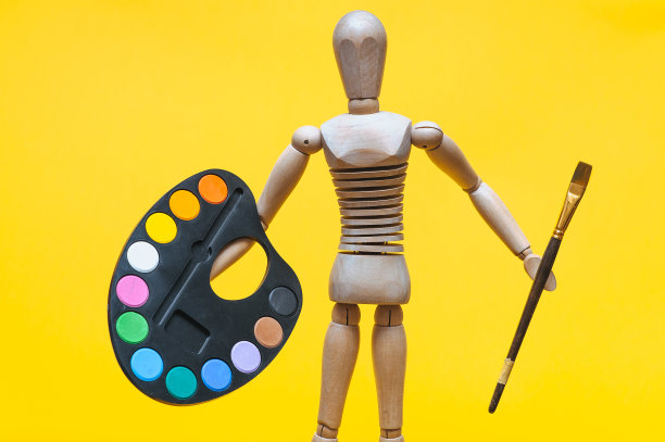 一个木头人手里拿着调色板和画笔，背景是黄色的。艺术家和创意概念。图片下载