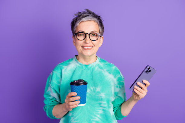 可爱的退休女士的照片肖像手持设备喝咖啡穿着时尚的绿色服装孤立在紫色的背景图片下载