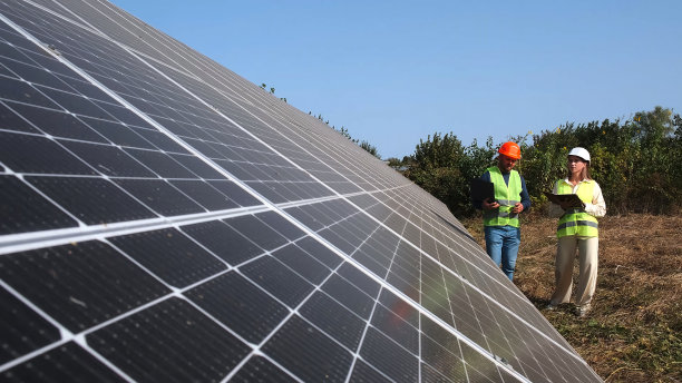 一男一女戴着安全帽，拿着笔记本电脑，正在太阳能电池板附近讨论工作任务。女环境工程师与投资者交谈。绿色电力理念。图片下载