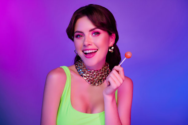 照片中有趣的女孩戴着金链珠宝，拿着美味的棒棒糖，孤立的多色霓虹灯背景图片下载
