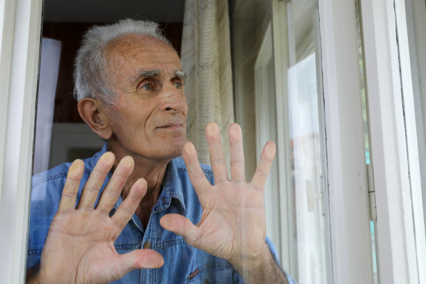 一位身穿蓝色牛仔衬衫的老人站在窗边，双手搭在窗前，从他的房间向外凝视。这张照片传达了一种内省和孤独的感觉。”图片下载