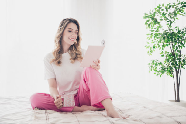 照片上的女孩正微笑着躺在床上抚摸头发，在白天白色的房间里看书图片下载