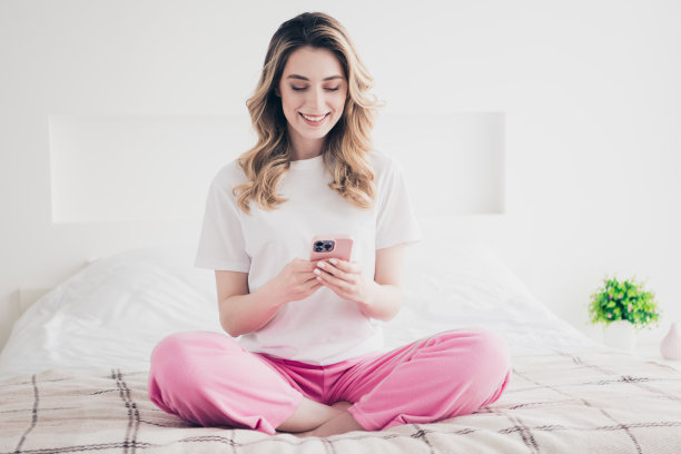 积极的女孩坐在床上的毯子，使用手机智能设备在灯光室内发短信的照片图片下载