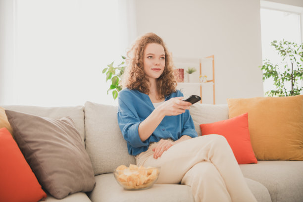 有吸引力的时髦年轻女子的照片坐在沙发上看电视吃薯片蓝色的衬衫搬家新公寓家庭室内室内图片下载