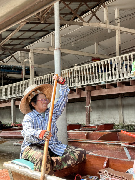 泰国Ratchaburi, Damnoen Saduak水上市场，船夫用大桨推着木船沿着运河行驶的特写照片，岸边的摊位和餐馆是前景的焦点图片下载