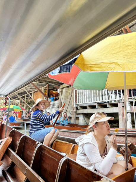 泰国Ratchaburi, Damnoen Saduak水上市场，船夫用大桨推着木船沿着运河行驶的特写照片，岸边的摊位和餐馆是前景的焦点图片下载