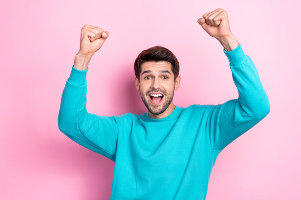 年轻时髦的学生鬃毛胡子的家伙穿着蓝色的套头衫拳头欢呼庆祝他的第一个盈利业务孤立在粉红色的背景图片下载