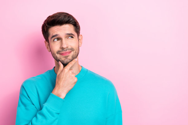照片中英俊的年轻人触摸下巴，看起来头脑好奇的空穿时髦的蓝色服装孤立在粉红色的背景图片下载