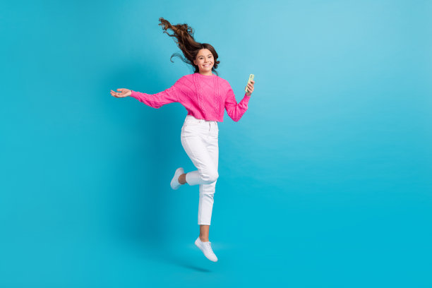 甜美漂亮女孩的全身照片穿着针织套衫跳高聊天苹果三星空空间孤立的蓝色背景图片下载