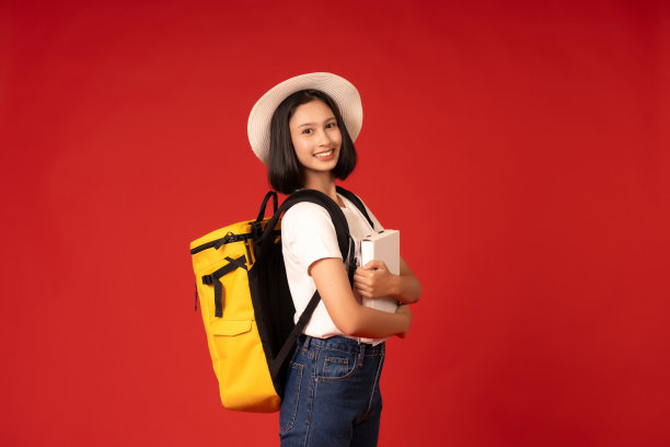 图为，在一所国际大学就读的亚洲女学生，在红色背景的摄影室内，面朝一侧，穿着便服，戴着帽子，拿着书，拿着包。图片下载