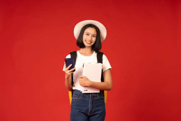 亚洲高中女生，辅导班和学校的主持人，用智能手机在线学习，穿着休闲的衣服和帽子，拿着书，在红色背景的工作室拍照。图片下载