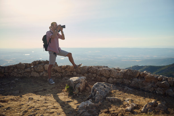 一名男子穿着休闲服，背着双肩包，在山顶上拍摄全景照片图片下载
