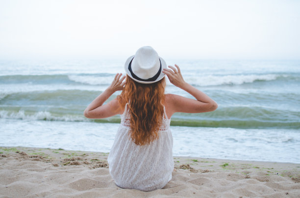 这是一幅宁静的照片，一个女人在海滩上，戴着夏天的帽子，穿着白色的裙子，享受着海景，在她放松的假期里，沐浴在海边的平静和宁静中图片下载