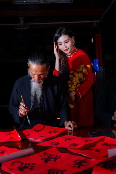 越南女孩穿着红色的敖黛服装和老学者在中国农历新年写春联。春联是中国春节期间的一项流行传统。照片中的文字意味着新年快乐，平平安安。图片下载