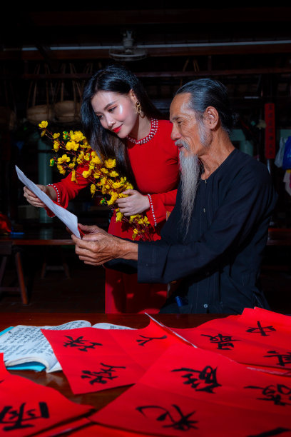 越南女孩穿着红色的敖黛服装和老学者在中国农历新年写春联。春联是中国春节期间的一项流行传统。照片中的文字意味着新年快乐，平平安安。图片下载