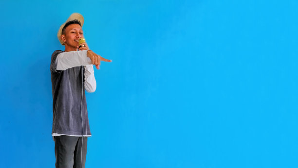 年轻富有表现力的歌手，快乐的男人，穿着灰色t恤，戴着眼镜和帽子，对着麦克风张开双手唱歌，在朴素的浅蓝色背景的工作室里。人们的生活方式观念图片下载