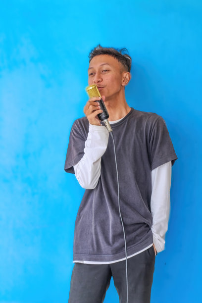 年轻富有表现力的歌手，快乐的男人，穿着灰色t恤，戴着眼镜和帽子，对着麦克风张开双手唱歌，在朴素的浅蓝色背景的工作室里。人们的生活方式观念图片下载