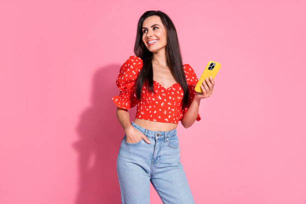 照片肖像的黑发西班牙年轻微笑的女士在明亮的红色时尚衬衫与智能手机看网站孤立的粉红色背景图片下载