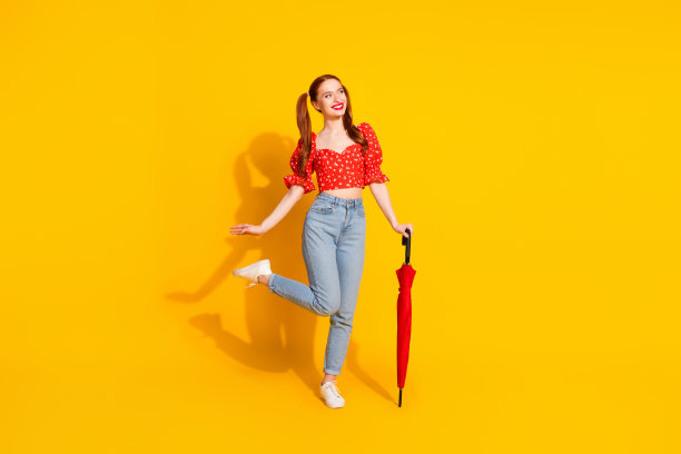 在春天的雨天里，穿着红色衬衫和牛仔裤的年轻女士手持阳伞的全身照片被孤立在黄色的背景上图片下载